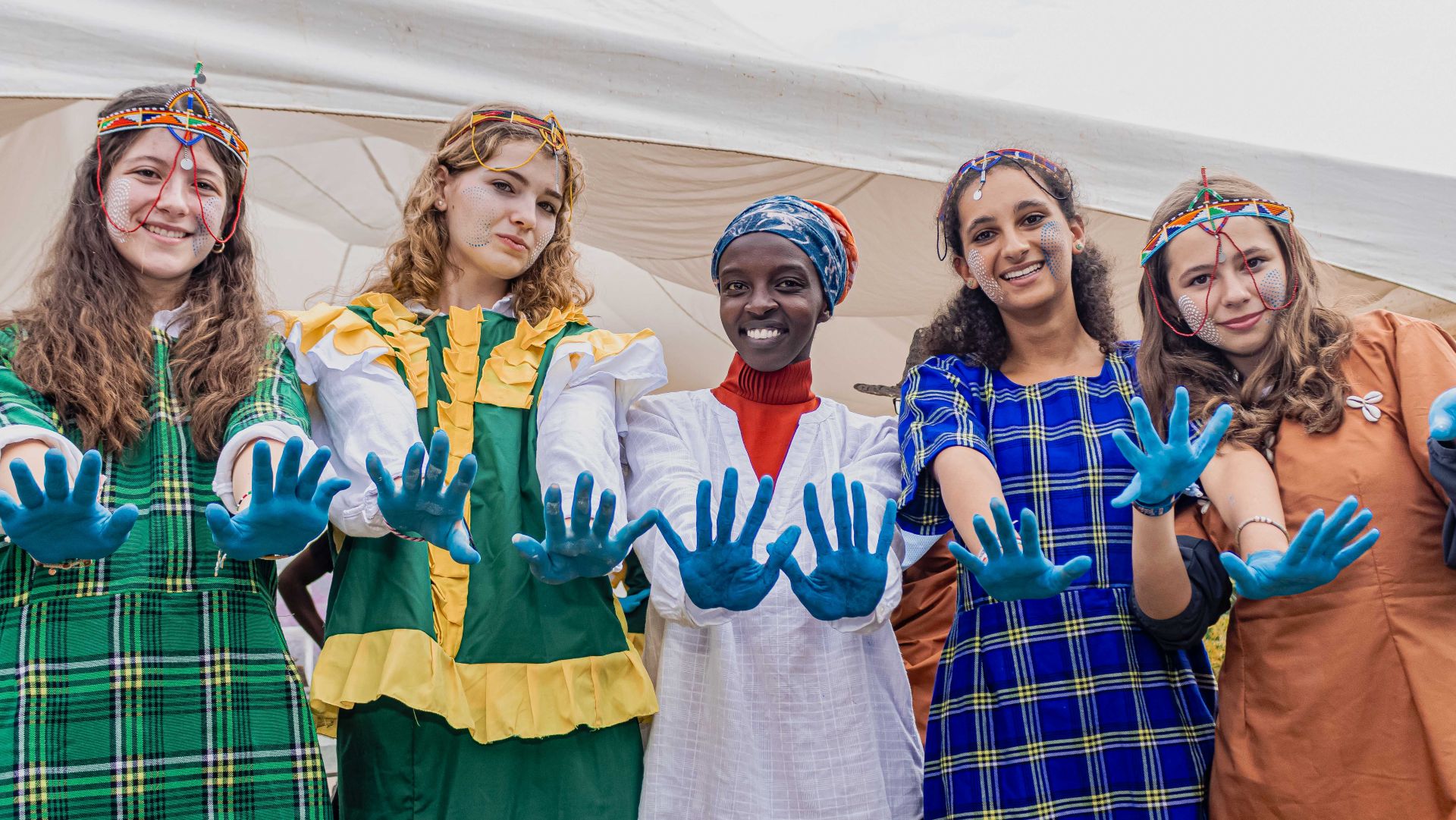 The image depicts a group of five young women standing in front of a white tent. They are dressed in vibrant outfits, suggesting a celebration or cultural event. Each woman is holding out her hands, which are covered in blue paint, towards the camera, creating an engaging scene. The event is taking place in Kenya.