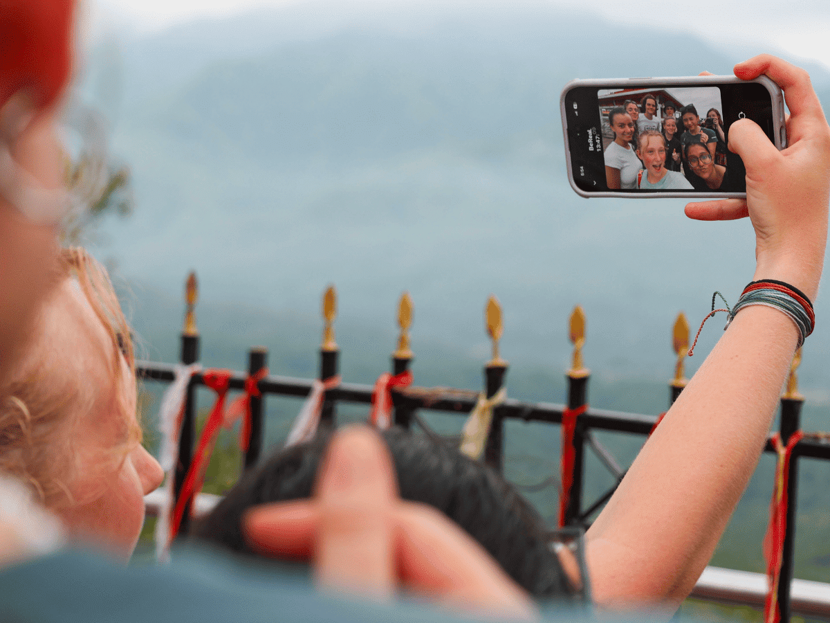 An over-the-shoulder view of a teenager holding up a smartphone to take a group selfie using the BeReal app. The phone screen displays a bright group photo of smiling teens. In the blurry foreground, the backs of the teenagers' heads are visible, and in the background, a black metal fence with gold tips stands before a vast, misty mountain landscape.