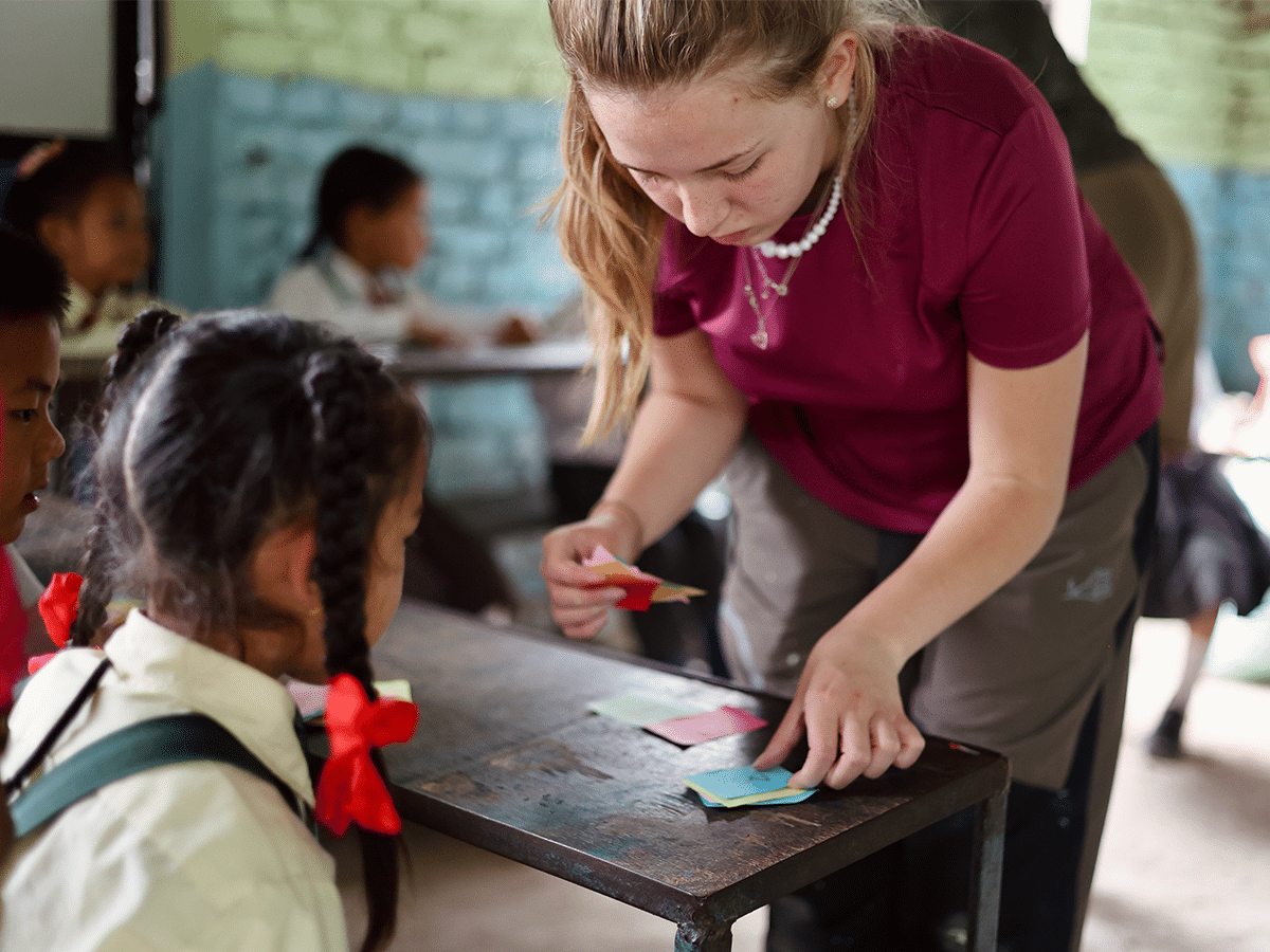 A young female volunteer leans over a wooden desk to teach a young student in a Nepalese classroom. The volunteer holds and arranges small, colorful paper cards on the table, while the girl, with her hair in braids tied with red ribbons, looks on attentively.