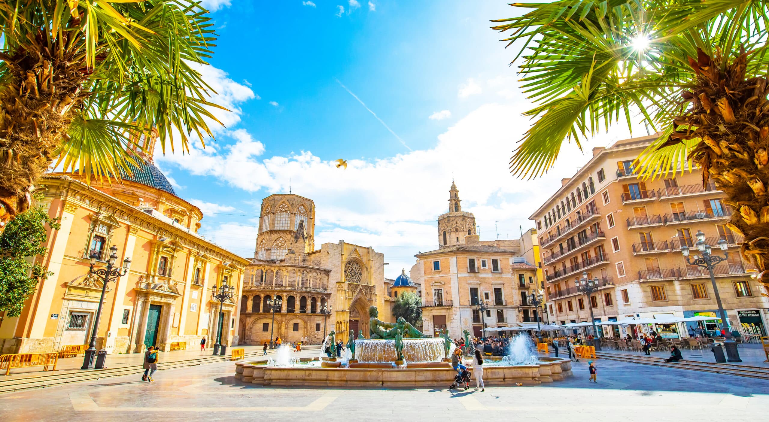 A sunlit view of Plaza de la Virgen in Valencia, Spain, framed by palm trees. A large fountain sits in the center of the stone square, surrounded by historic buildings, including the Valencia Cathedral and Basilica under a bright blue sky.